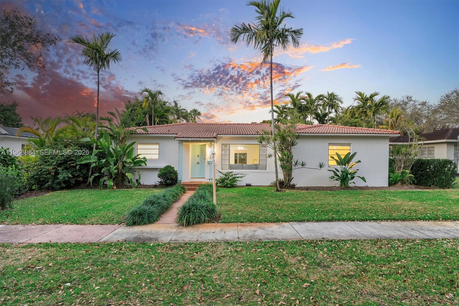 Single-story white home with a tile roof, palm trees, and tropical landscaping at dusk.