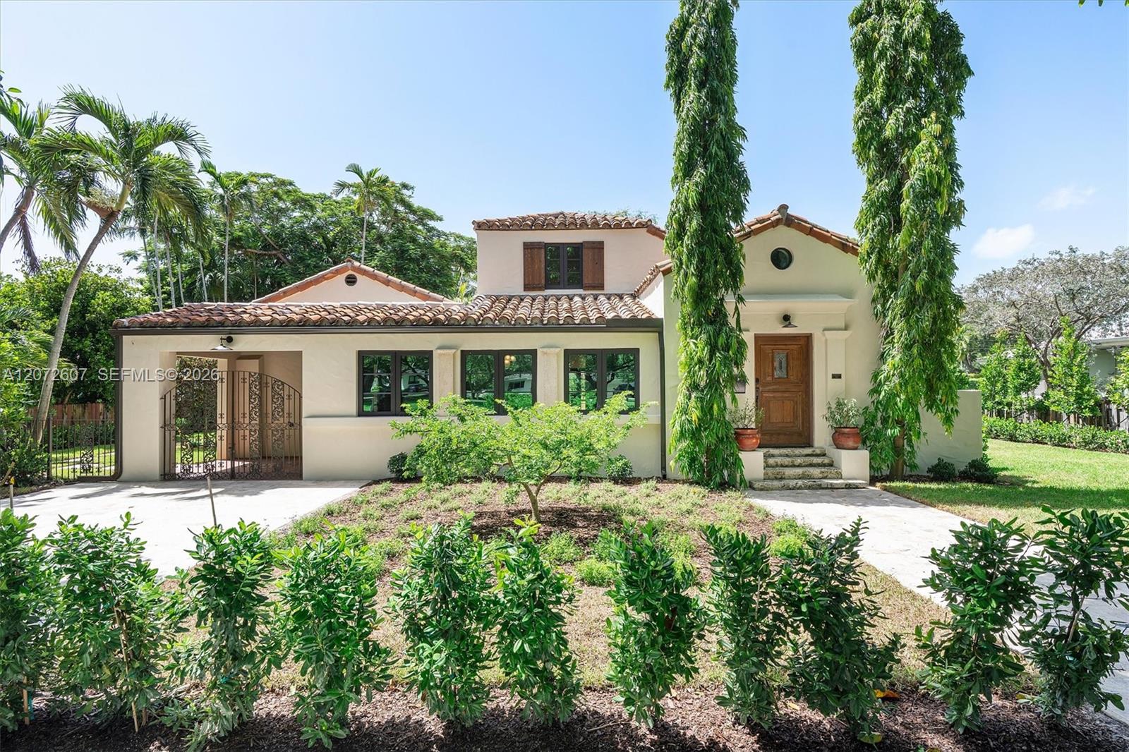 Cream stucco home with terracotta roof, gated carport, and tropical landscaping.