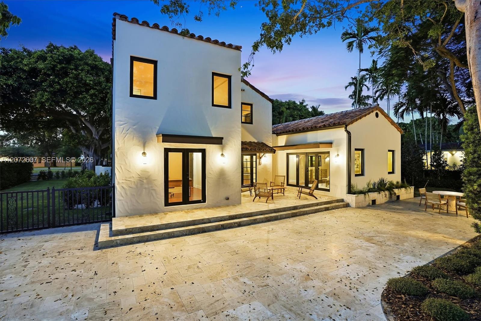 White stucco home with tiled roofs, warmly lit windows, and a spacious stone patio at dusk.