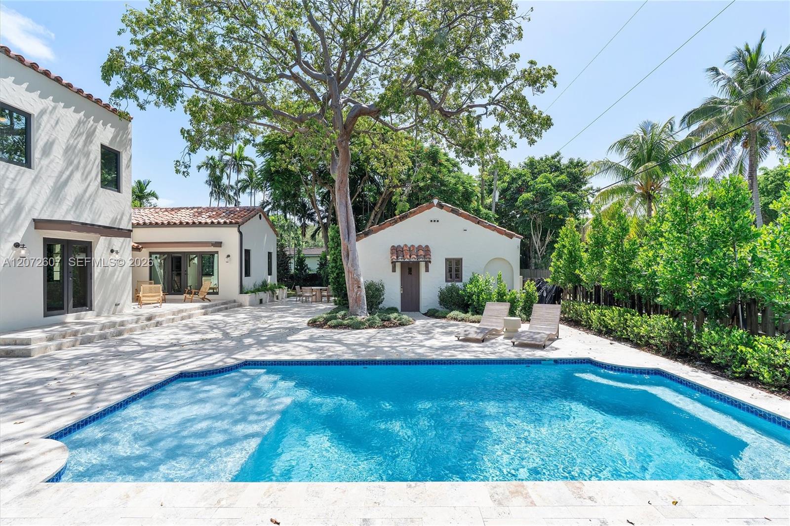 Backyard pool beside a white stucco home, with a tiled patio and mature tropical landscaping.