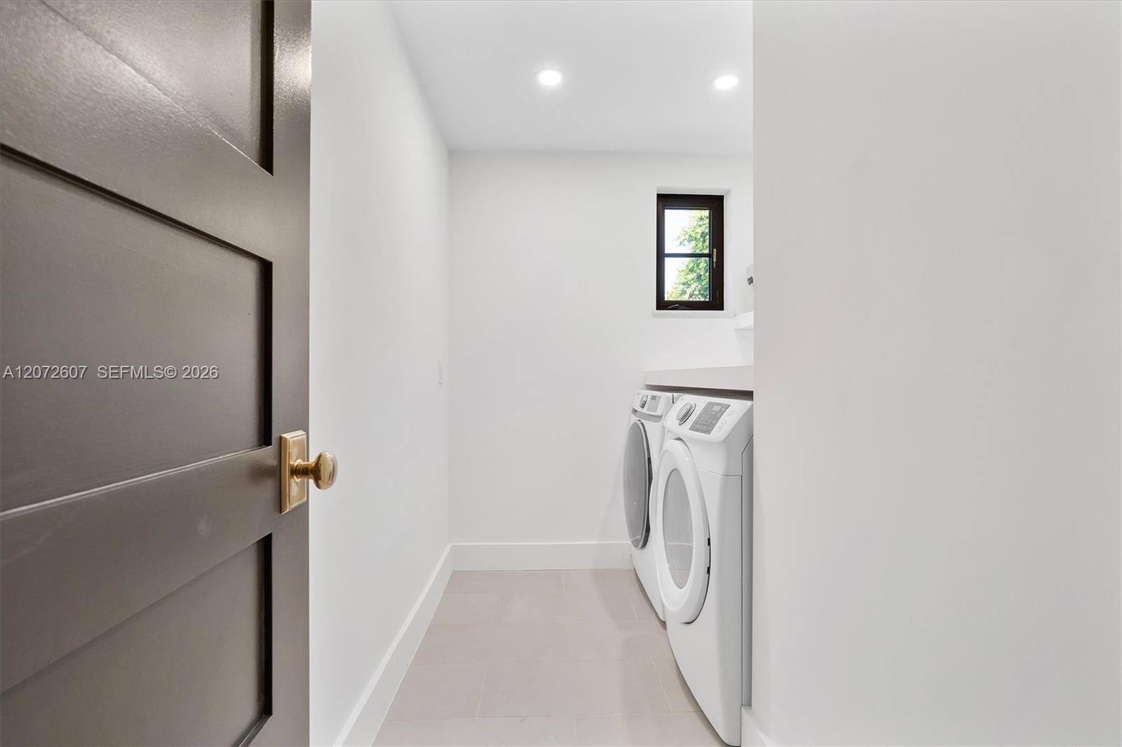 Laundry room with white washer and dryer beneath a countertop and a small high window.
