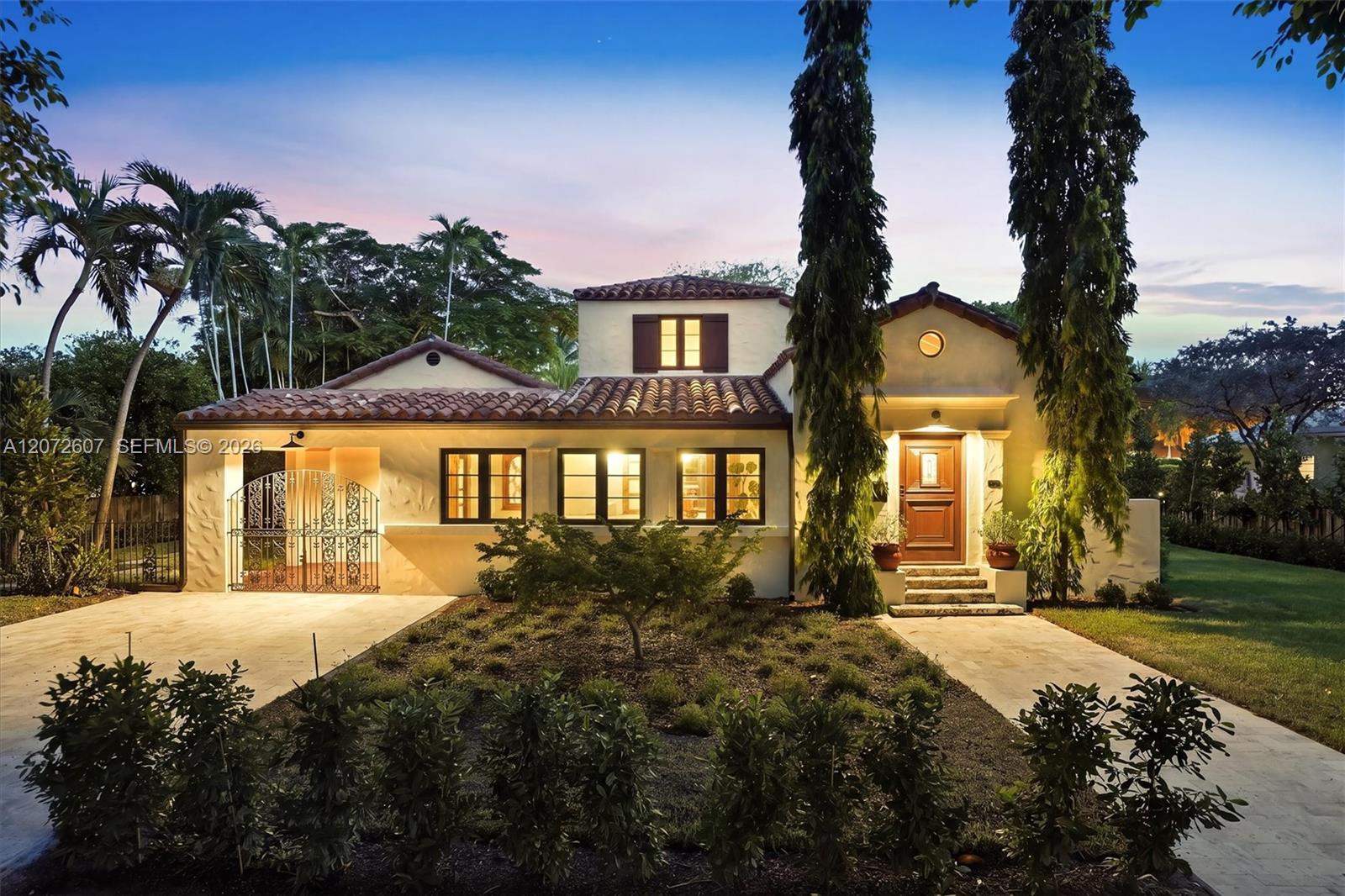 Two-story stucco home with a tile roof, illuminated windows, tall palms, and an ornamental gate at dusk.