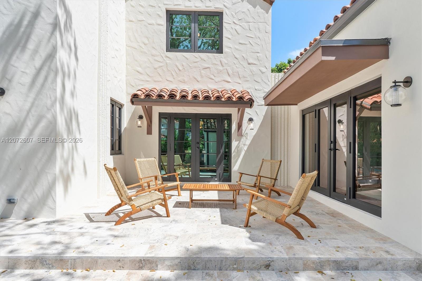 Stone patio courtyard with woven lounge chairs, a wooden table, and dark-framed glass doors.