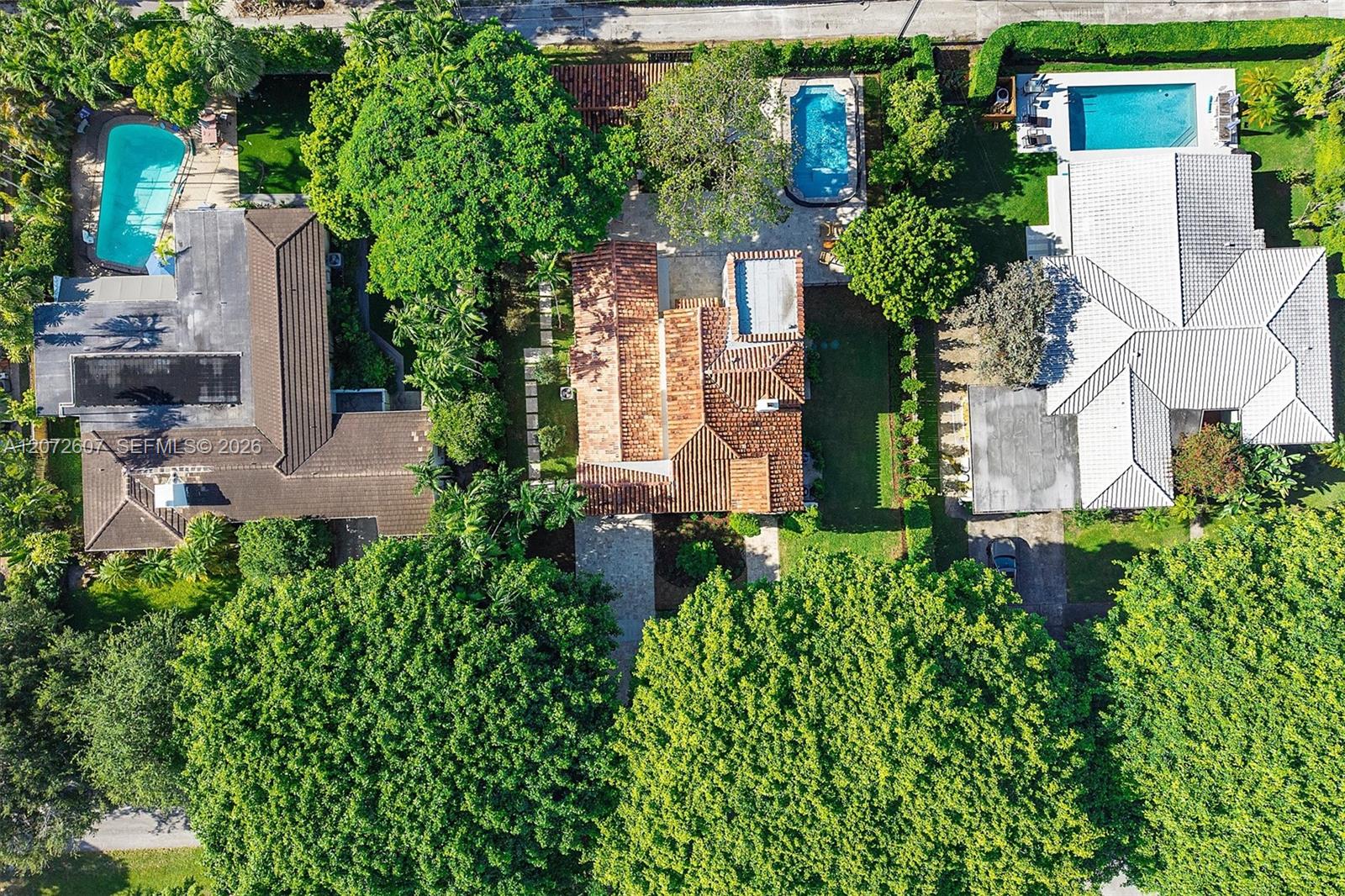 Aerial view of a terracotta-roofed home surrounded by tropical trees, with neighboring pools and houses.