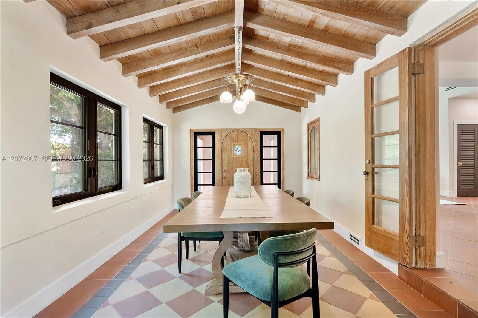 Dining room with a vaulted wood-beam ceiling, colorful tile floor, and long table with green chairs.