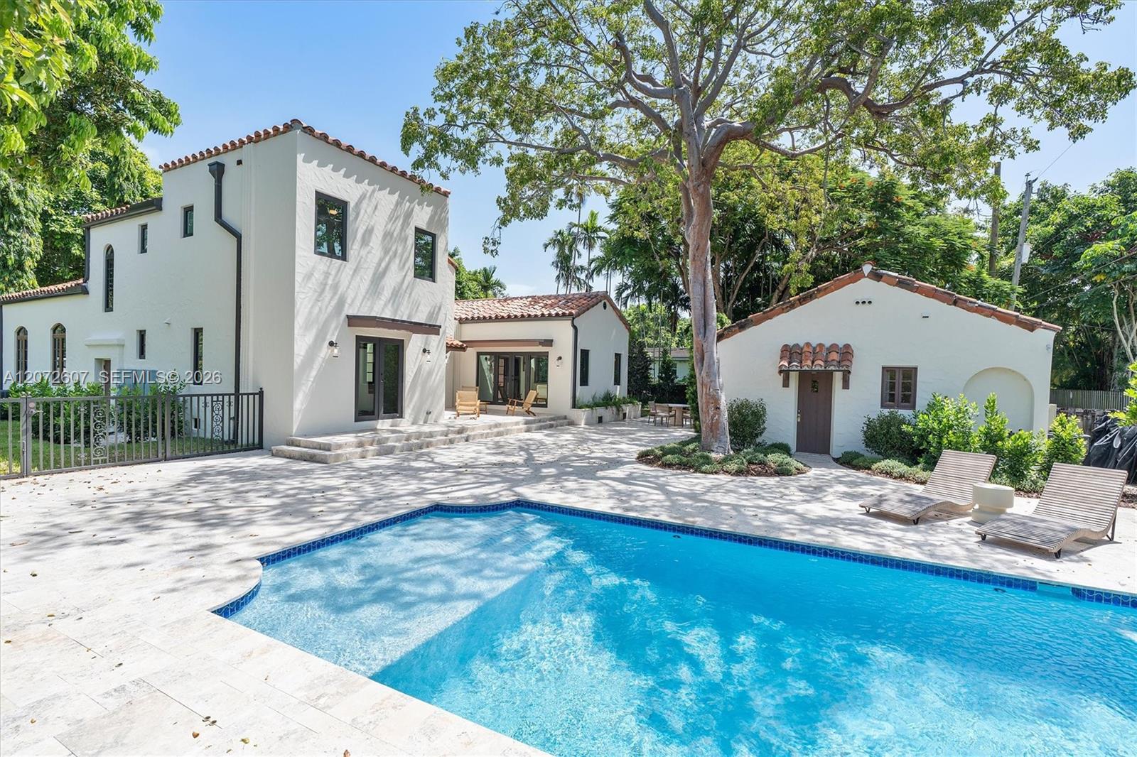 Backyard swimming pool beside a white Spanish-style home, shaded by a large tree.