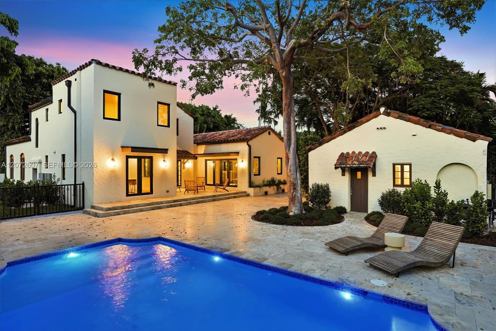 Spanish-style home and illuminated swimming pool beside a paved patio at dusk.