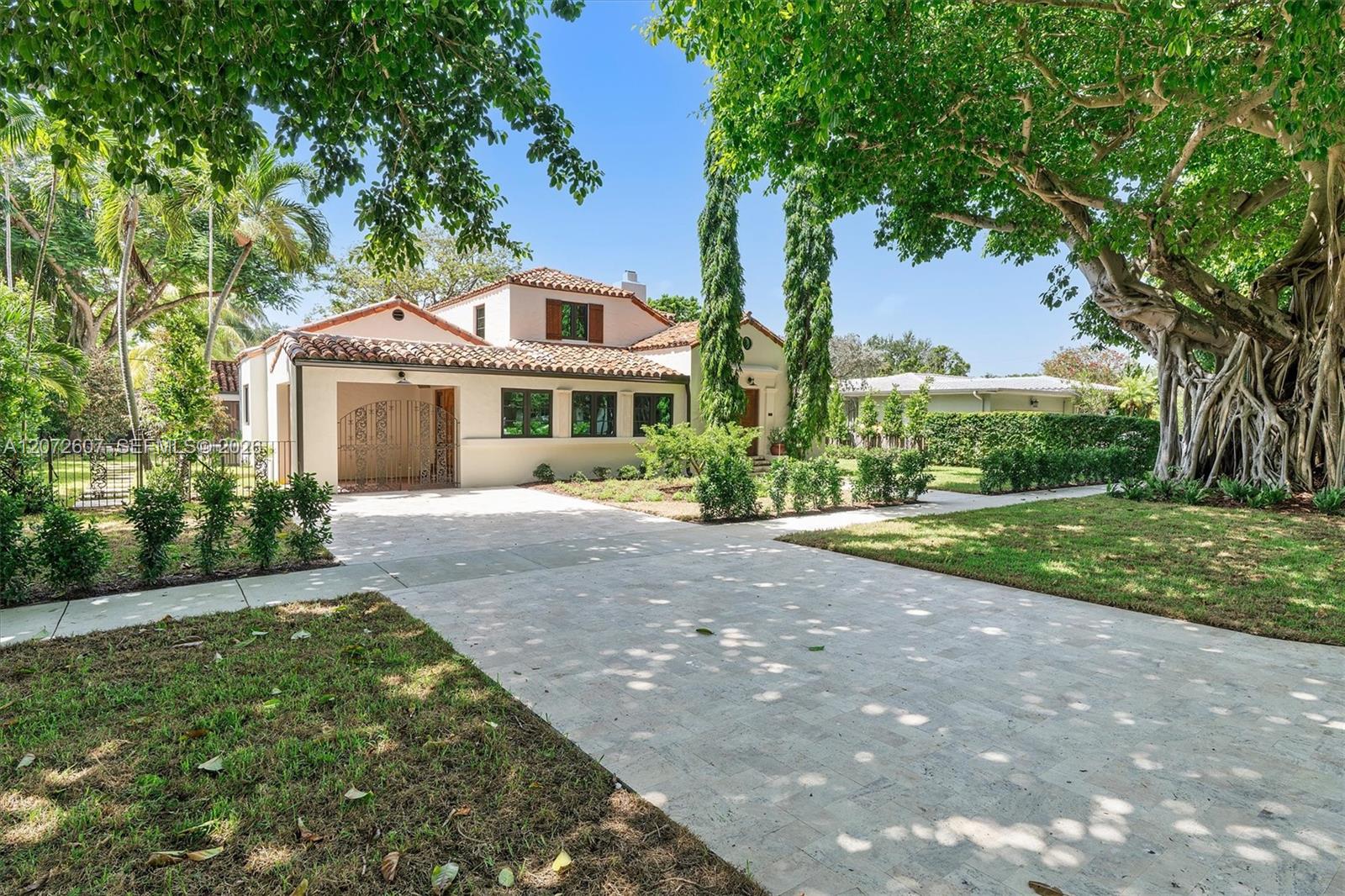 Stucco home with a terracotta tile roof, broad paved driveway, and mature tropical trees.