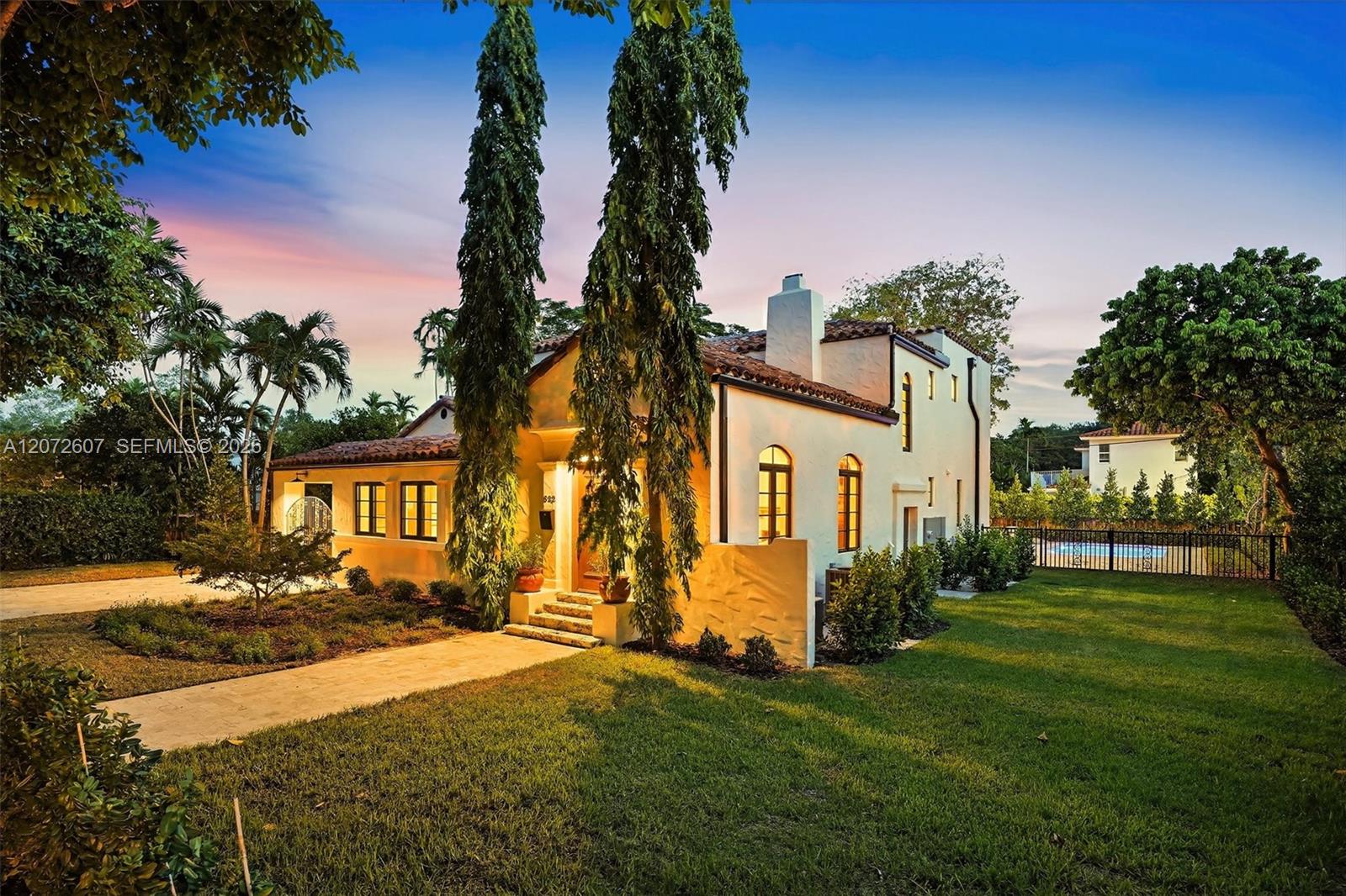 White stucco home with a tile roof, tall palms, landscaped lawn, and fenced swimming pool at sunset.