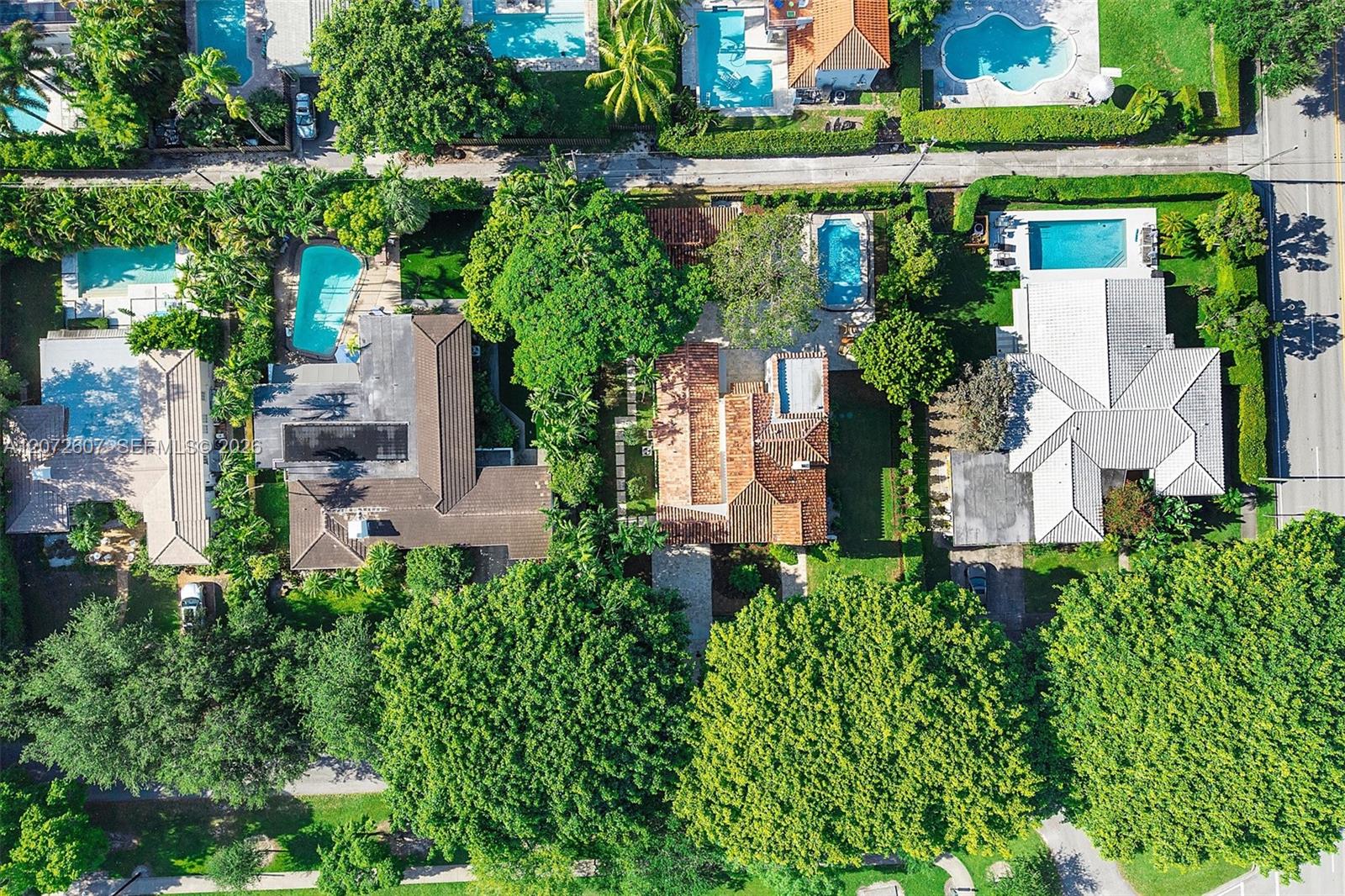 Aerial view of a terracotta-roofed home surrounded by trees, neighboring houses, and swimming pools.
