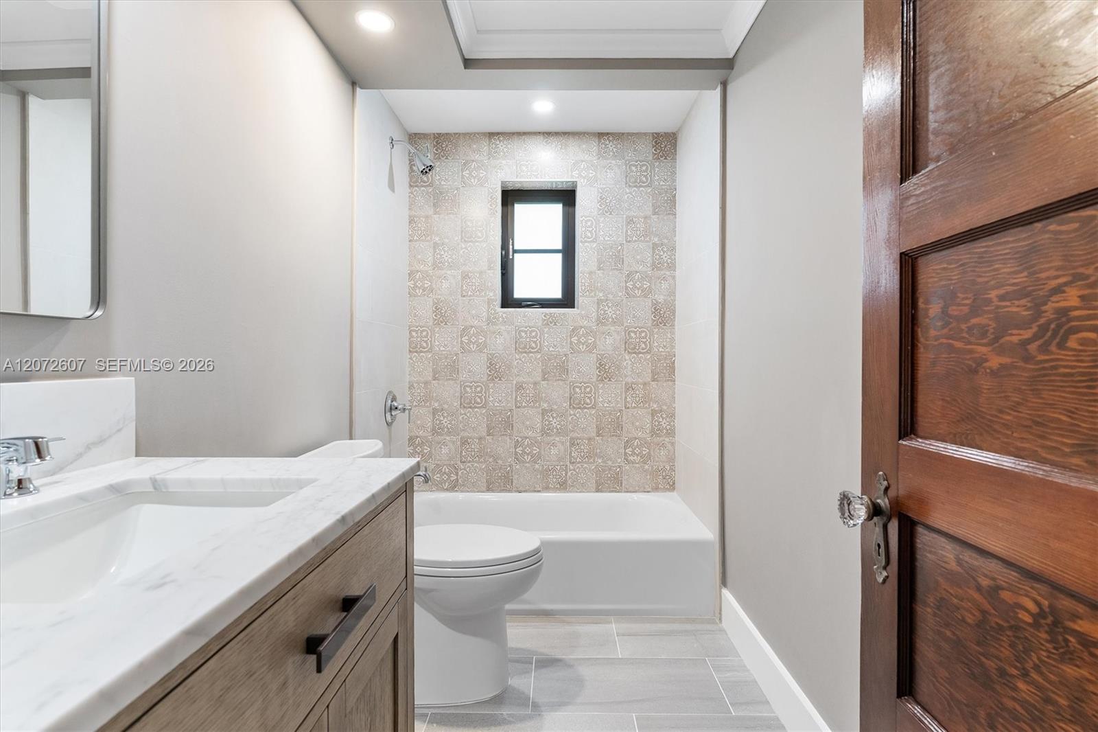 Bathroom with a marble-look vanity and bathtub backed by patterned tile and a small window.
