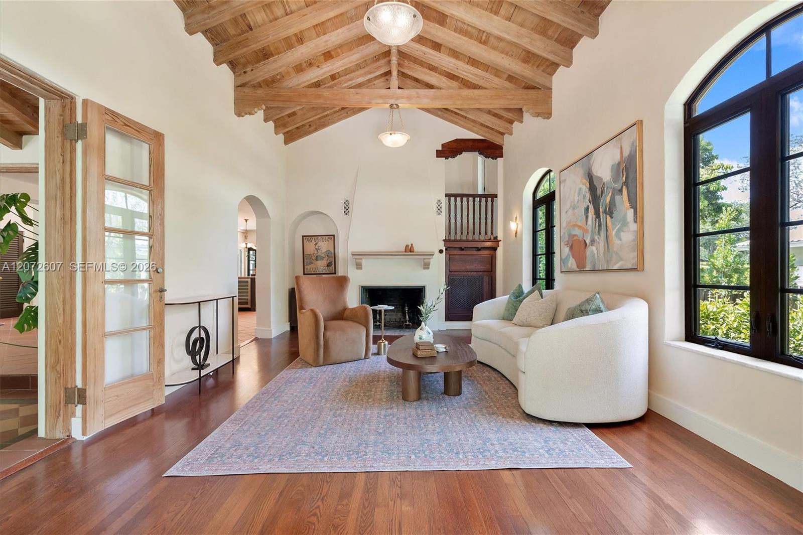 Living room with exposed wood beams, a fireplace, and tall arched windows.