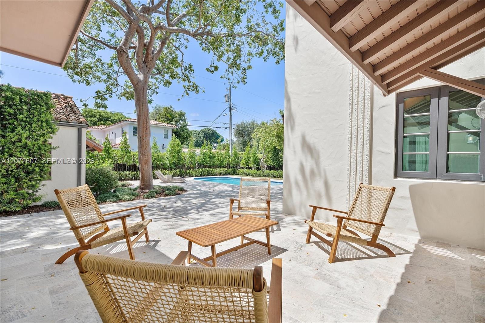 Covered stone patio with woven lounge chairs and a wood table overlooking the backyard pool.
