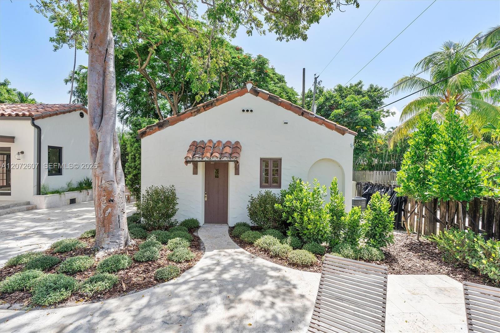 White stucco home with terracotta roof, framed by tropical trees and a landscaped paved courtyard.