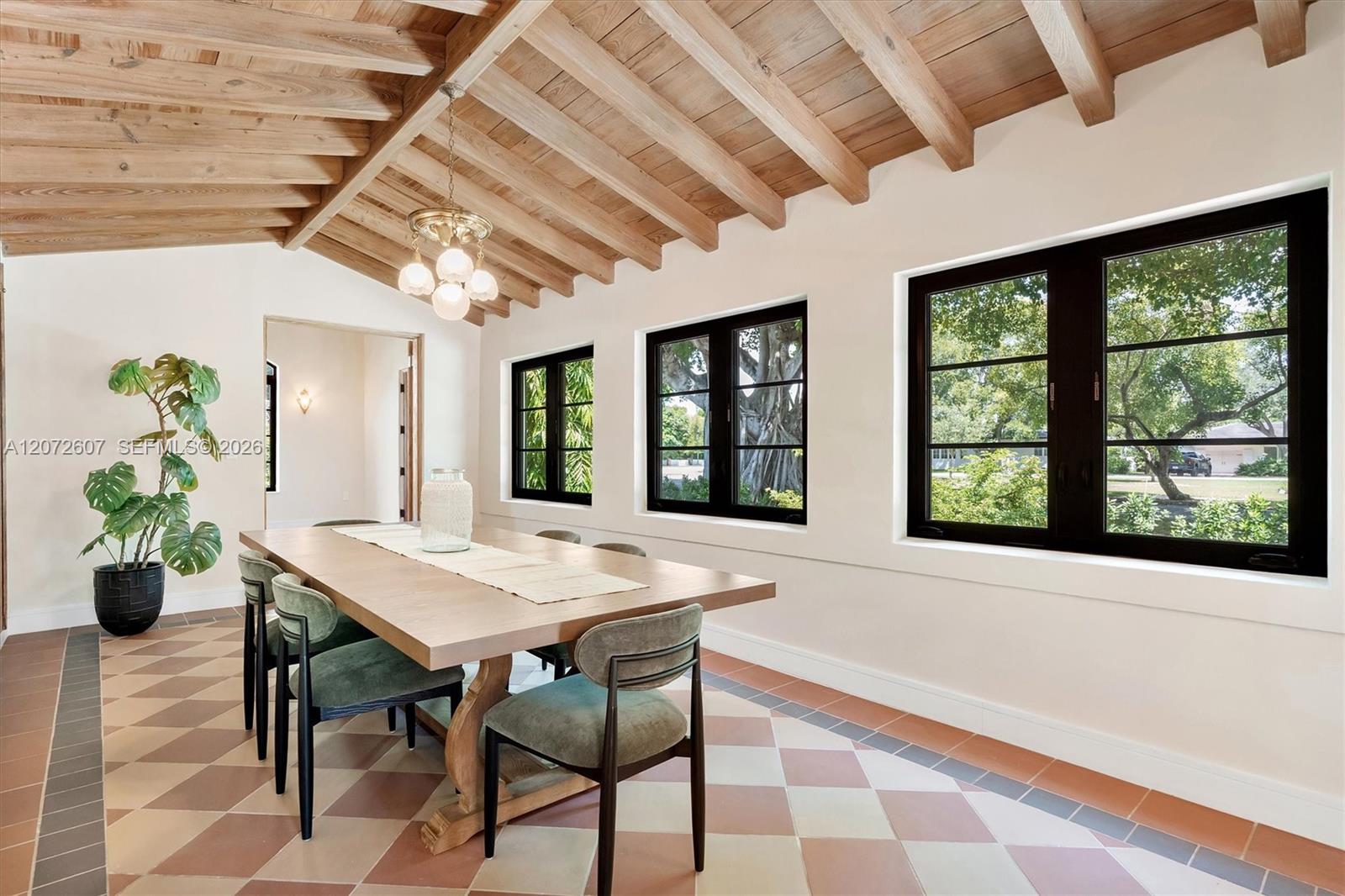 Dining room with exposed wood beams, patterned tile flooring, and black-framed windows overlooking trees.
