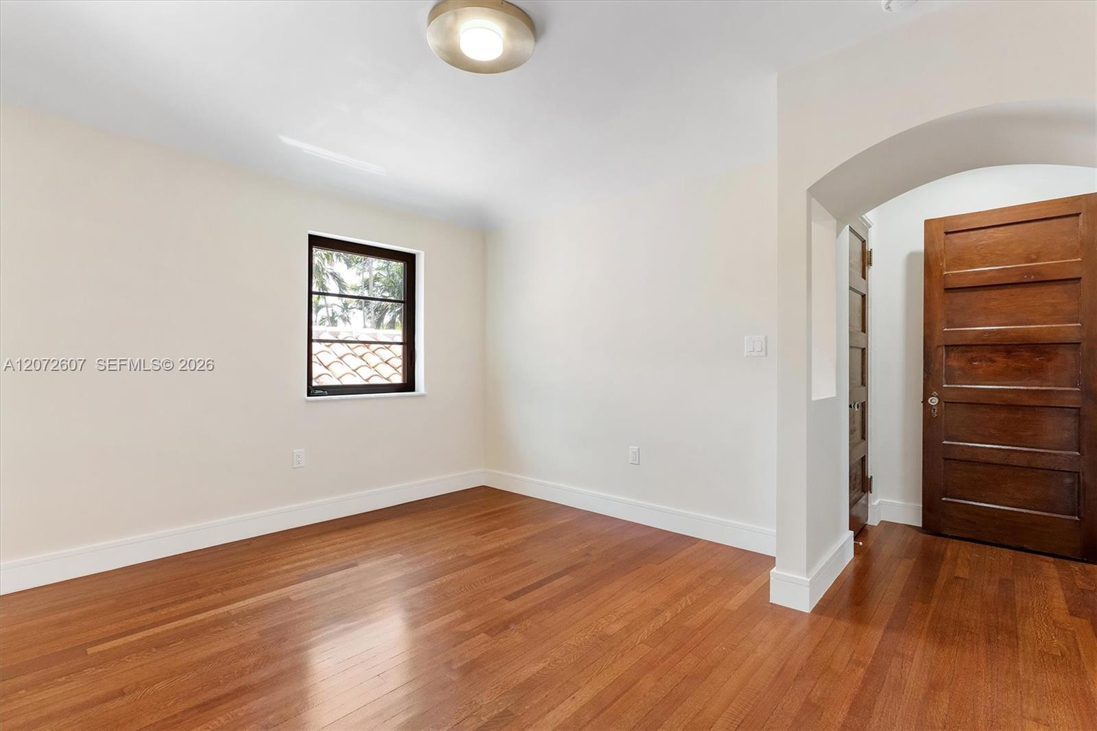 Empty room with polished hardwood floors, a small window, and an arched doorway beside a dark wood door.