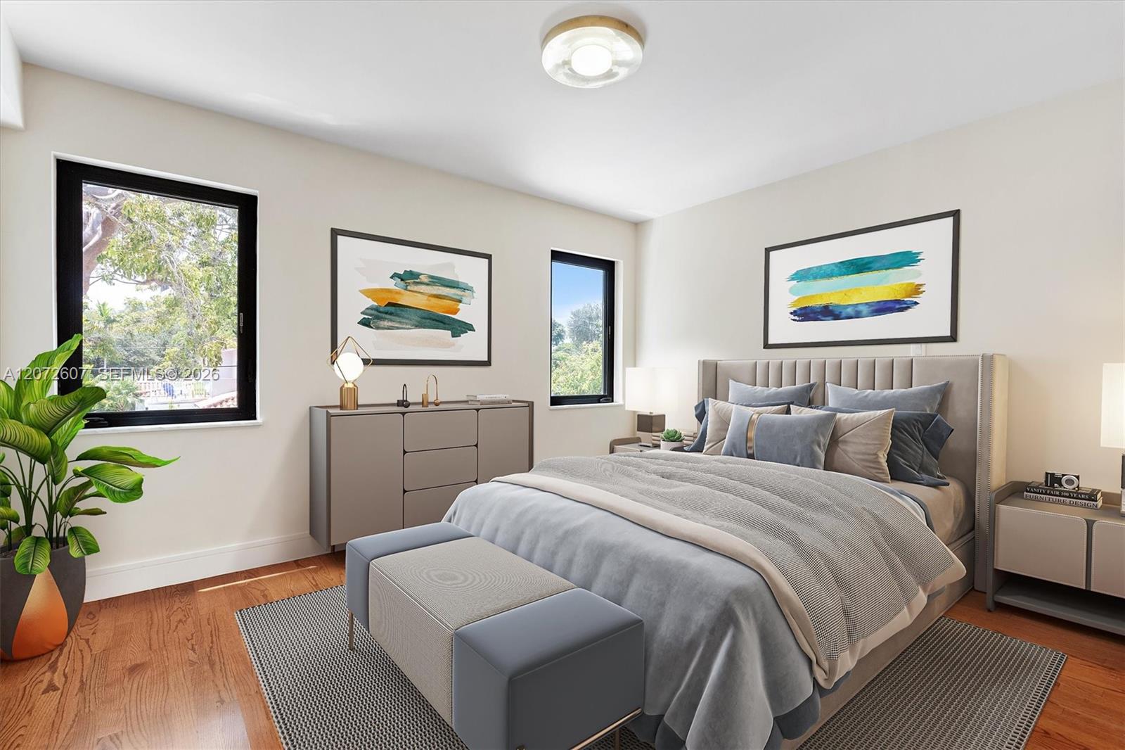 Staged bedroom with a gray upholstered bed, hardwood floors, and two tall windows overlooking greenery.