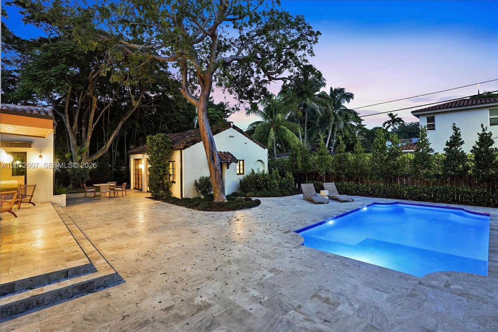 Backyard swimming pool beside a paved patio, lounge chairs, and a white stucco home at dusk.