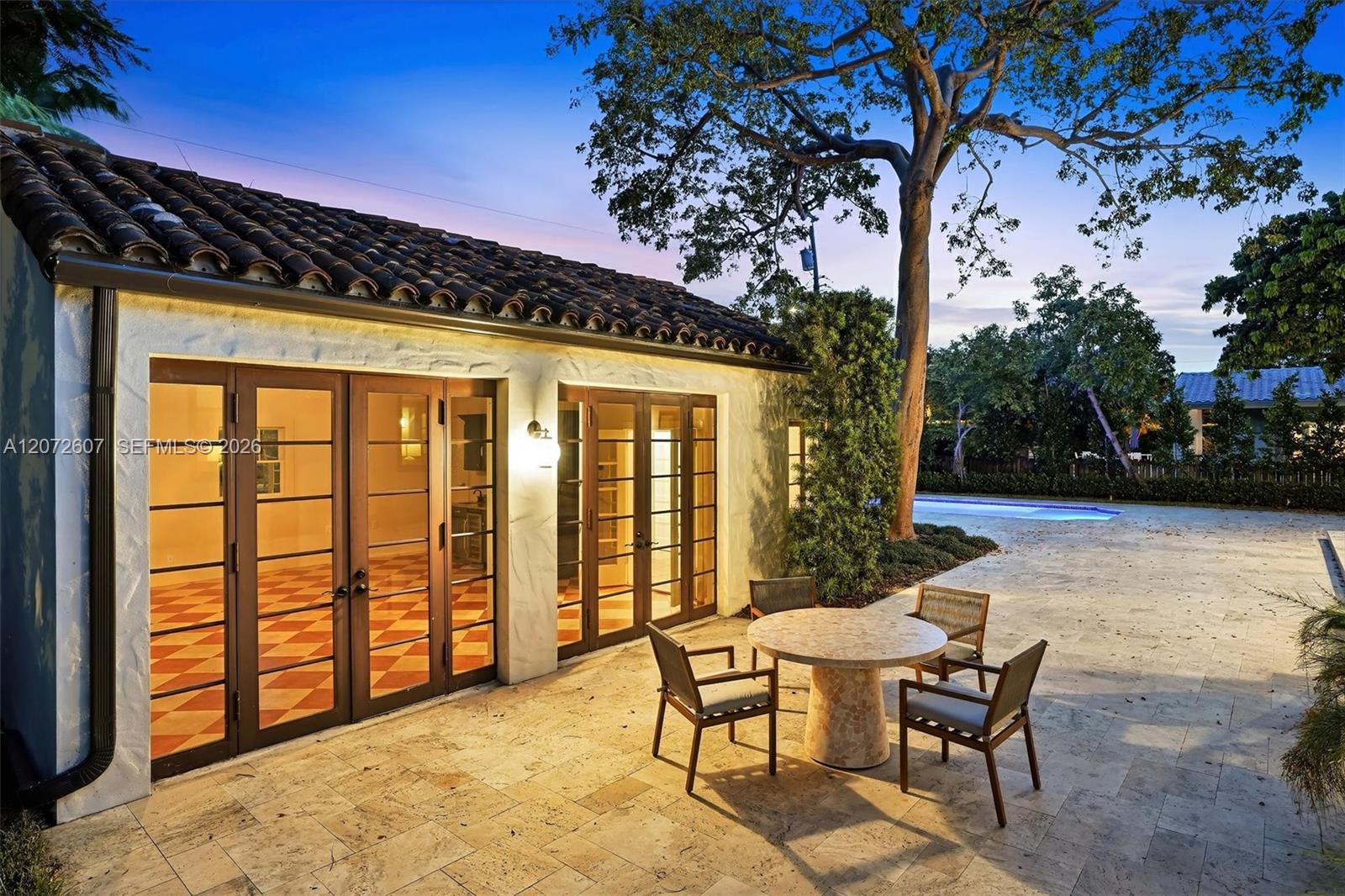 Home exterior with illuminated French doors, a stone patio, outdoor seating, and a pool beneath a large tree.