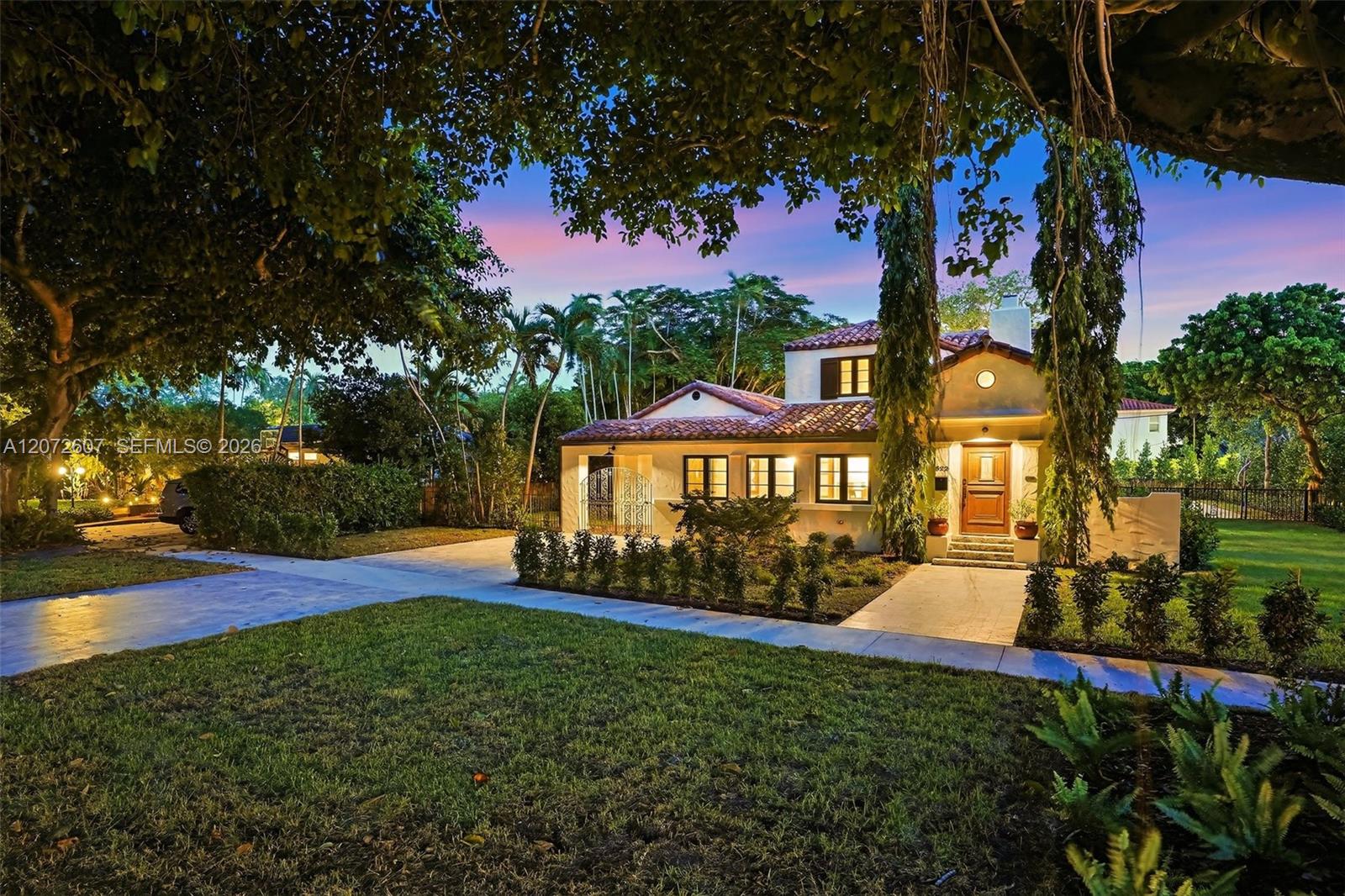 Stucco home with a red tile roof and lit windows framed by mature trees at dusk.