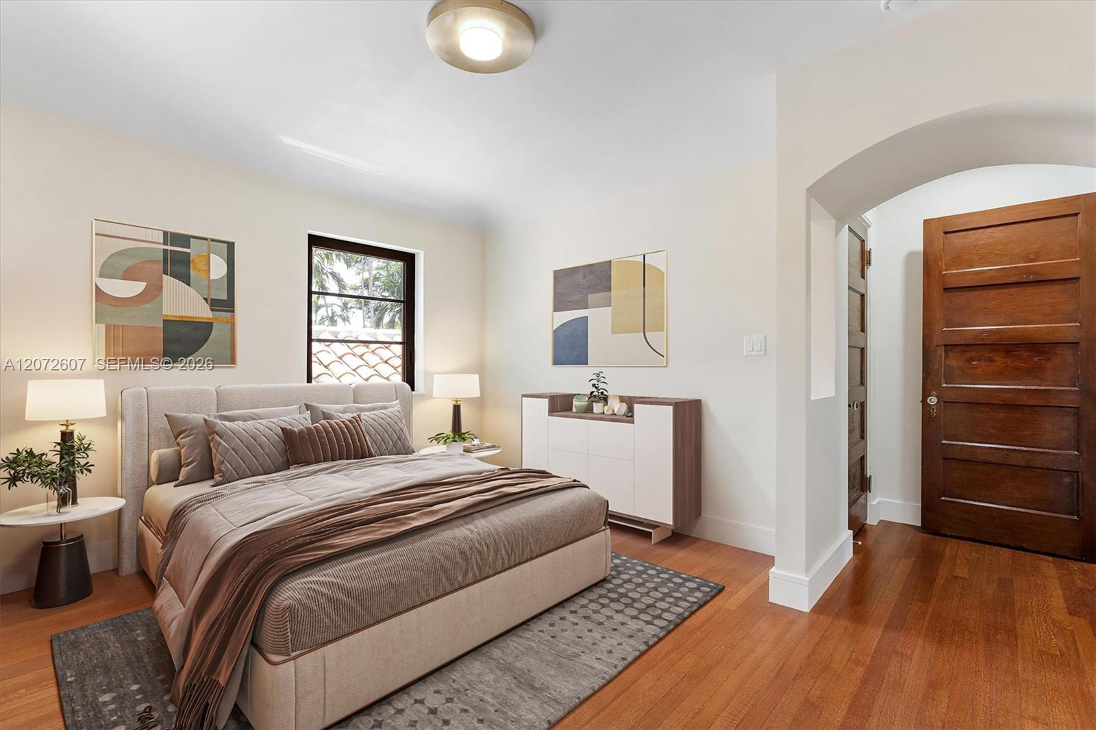 Staged bedroom with a large upholstered bed, abstract wall art, hardwood floors, and an arched doorway.