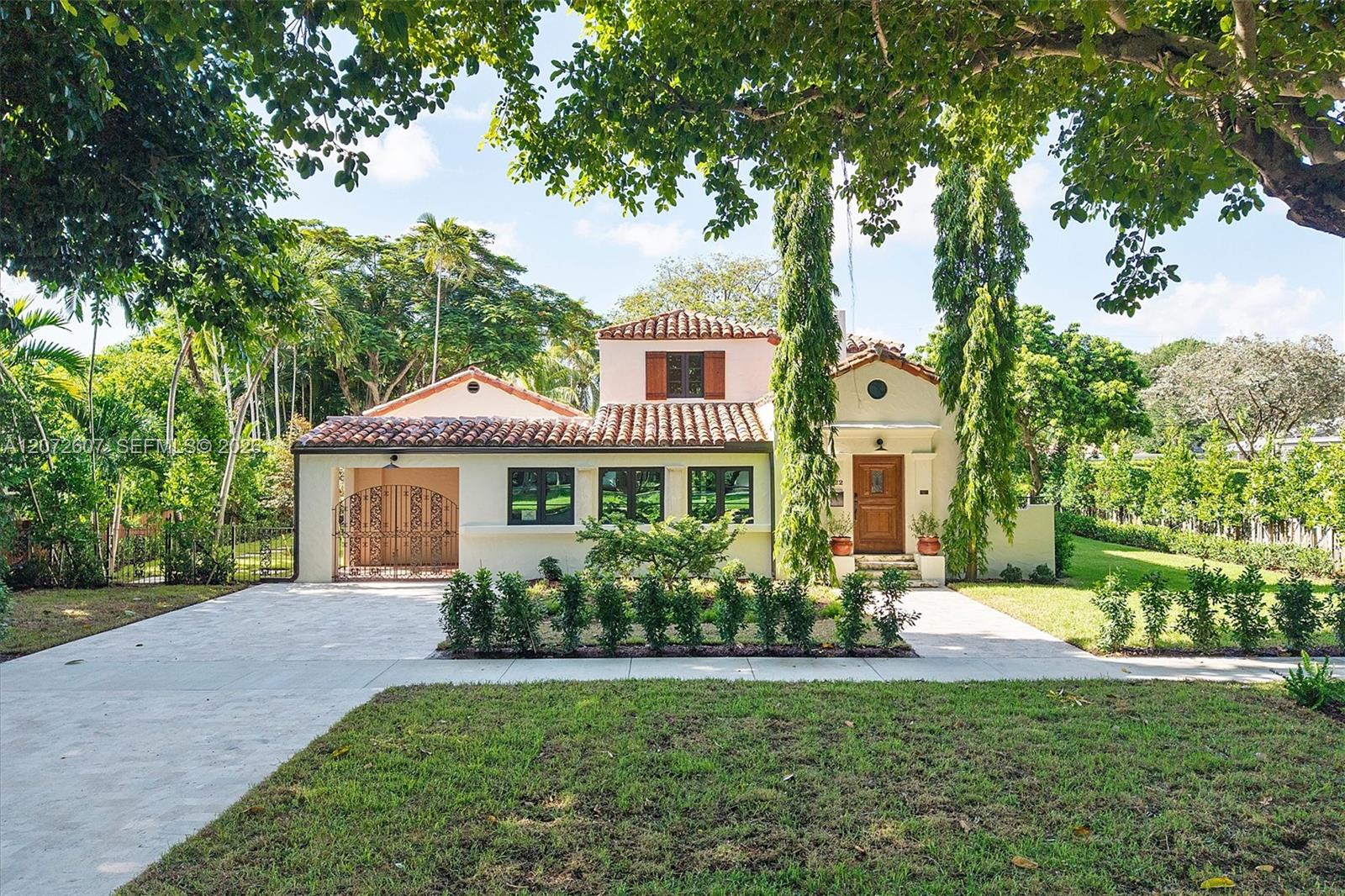 Stucco house with a terracotta tile roof, arched driveway gate, and mature tropical landscaping.