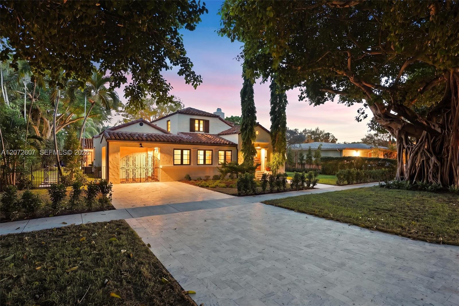 Two-story stucco home with a tiled roof and broad paved driveway, framed by mature trees at dusk.