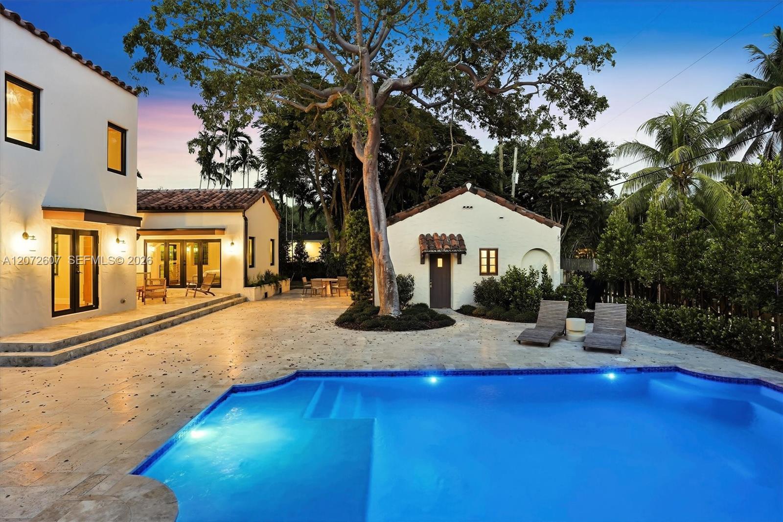 Backyard pool and stone patio beside a white stucco home, framed by tropical trees at dusk.
