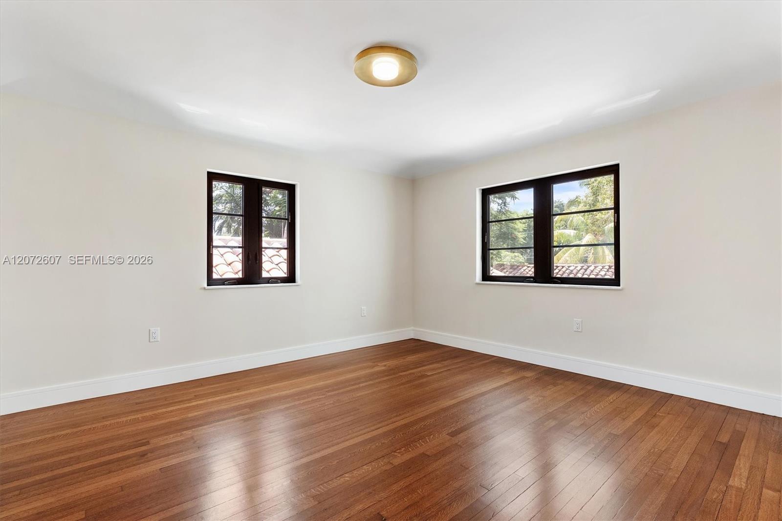Empty room with polished hardwood floors and two dark-framed windows overlooking trees and a tiled roof.