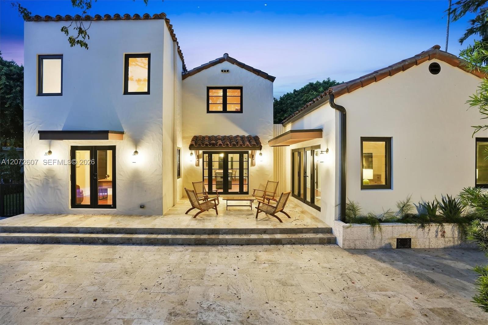White stucco home at dusk with a stone patio, tiled roofs, and a seating area.