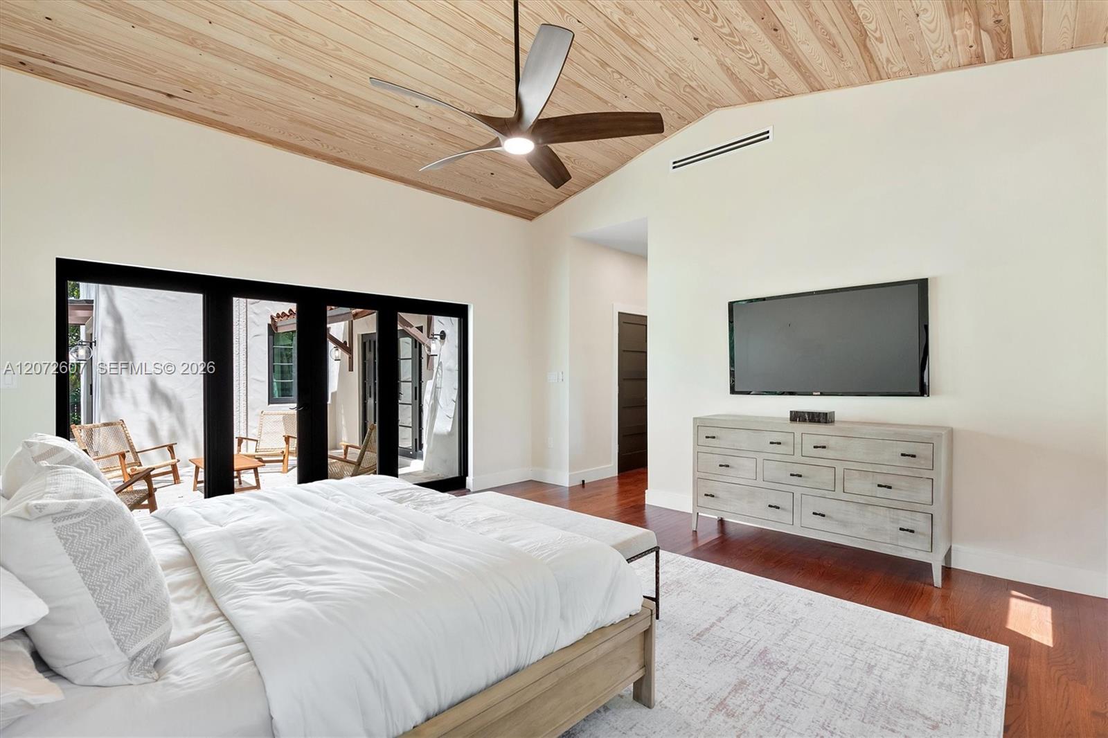 Bedroom with a white bed, wood-plank ceiling, and black-framed doors opening to an outdoor seating area.
