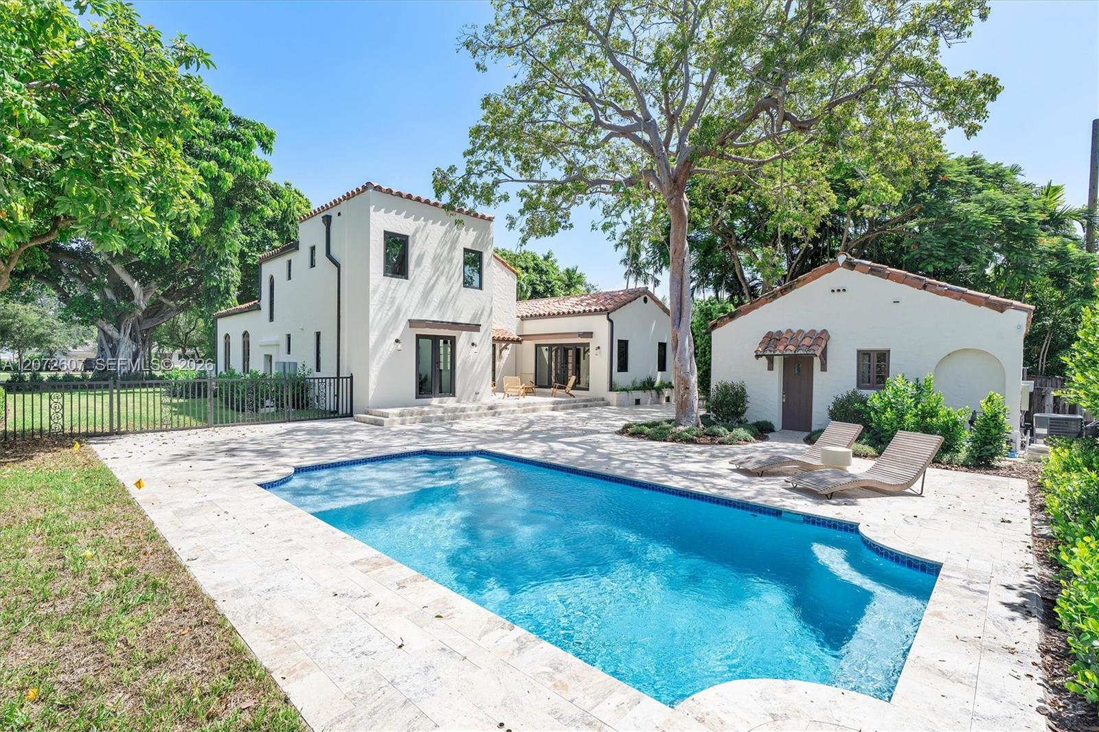 White Spanish-style home and detached outbuilding beside a swimming pool, with a stone patio and mature trees.