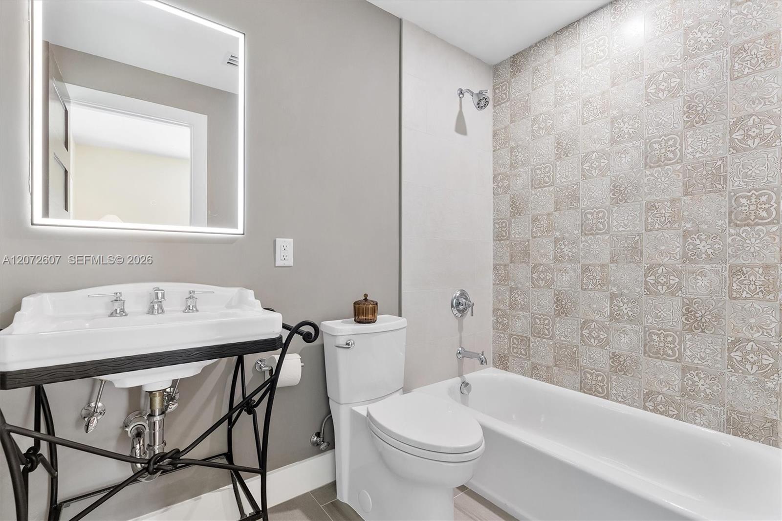 Bathroom with patterned gray shower tiles, a white tub and toilet, and a sink beneath an illuminated mirror.
