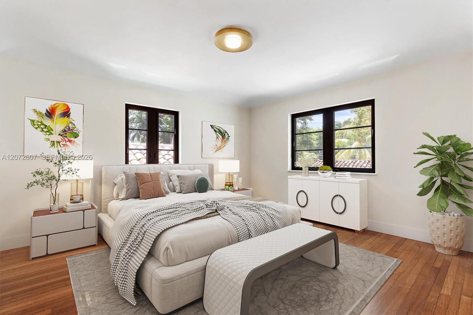Staged bedroom with a light upholstered bed, patterned throw, and dark-framed windows overlooking greenery.