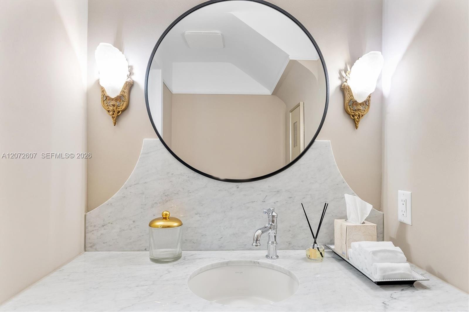 Bathroom vanity with a white marble counter, round mirror, and ornate gold sconces.