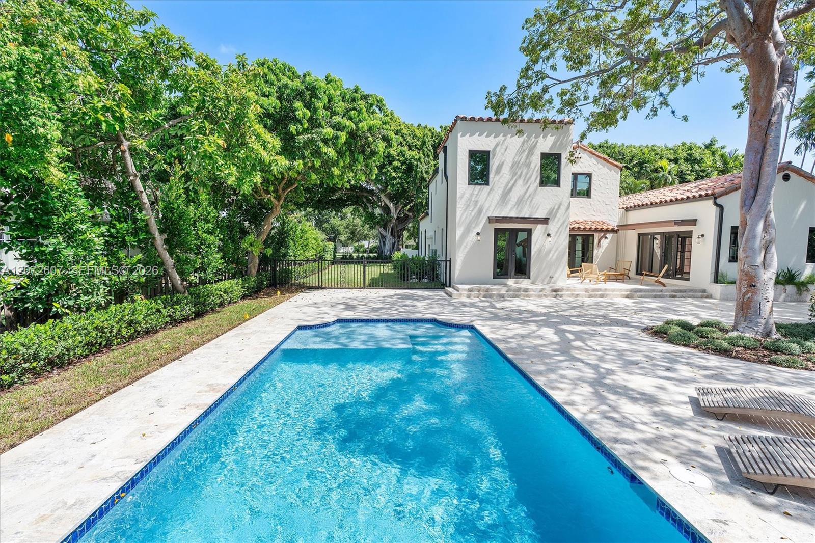 Backyard pool beside a white stucco house, with a stone patio and mature trees.