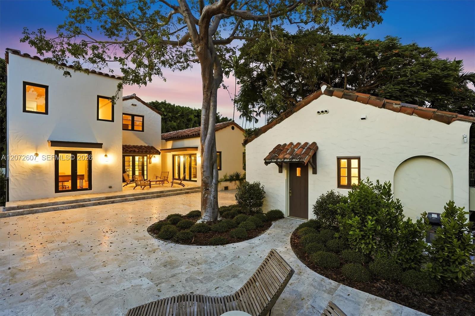 Spanish-style home with a tiled courtyard, mature tree, and warmly lit windows at dusk.
