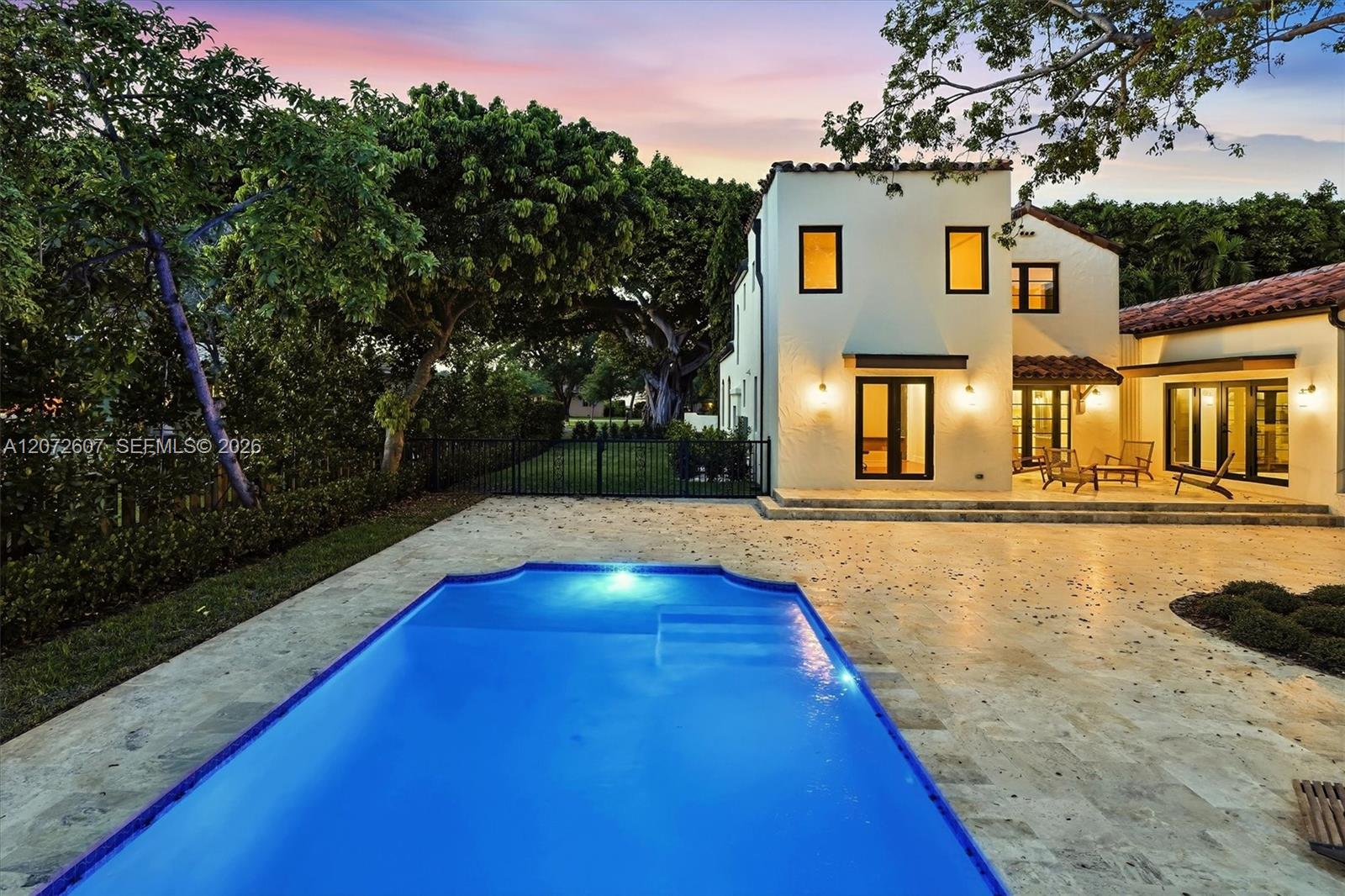 White stucco home and furnished patio beside a bright blue swimming pool at dusk.