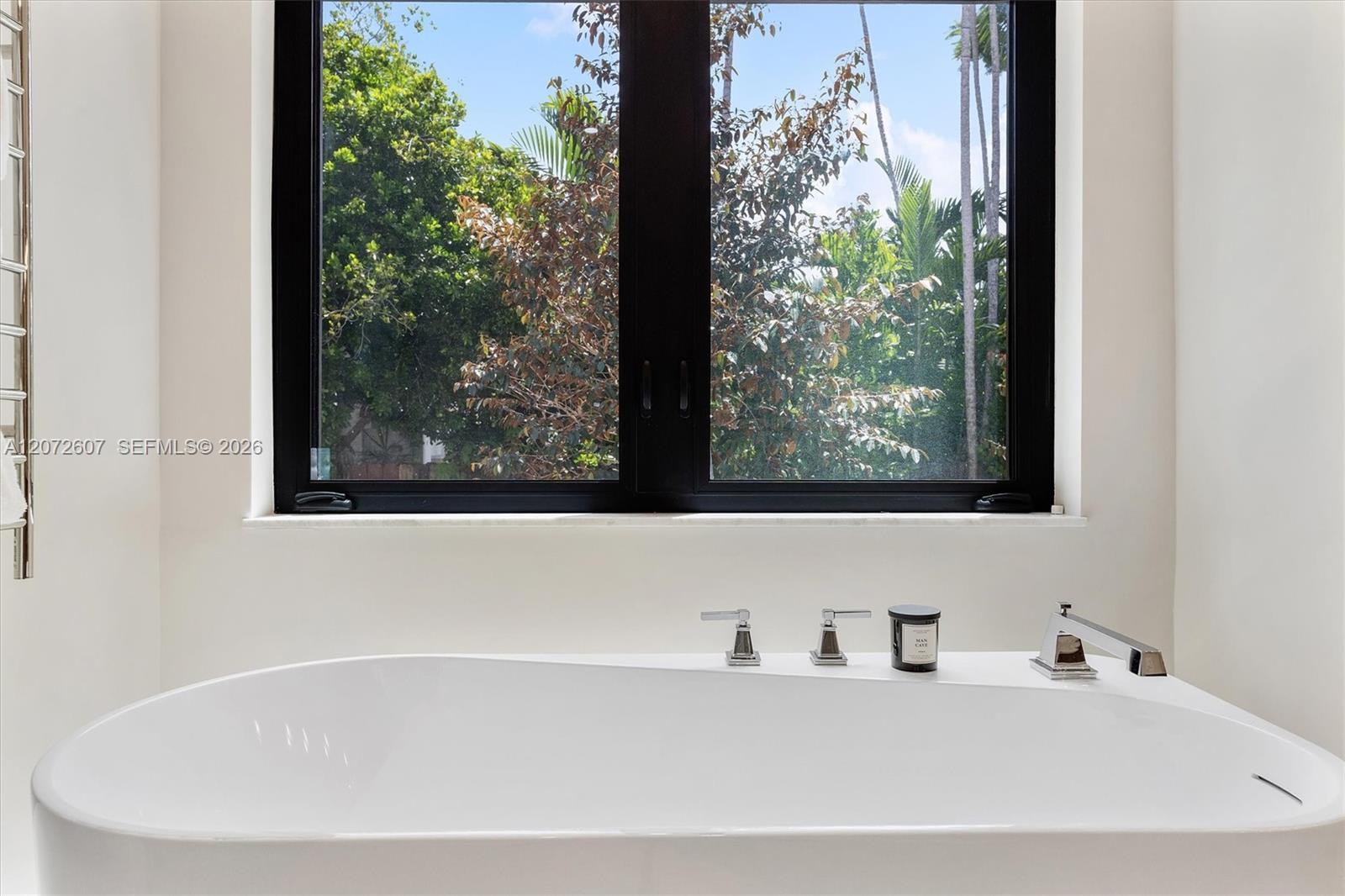 Freestanding bathtub beneath a large window with leafy trees outside.