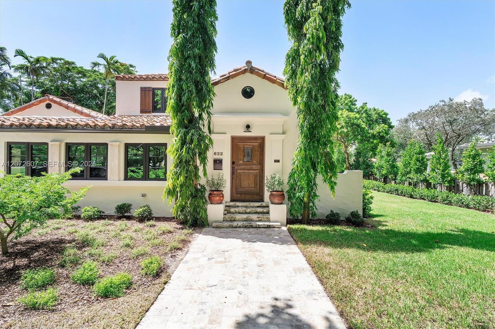 Cream stucco home with a tile roof, wooden front door, and stone walkway between tall trees.