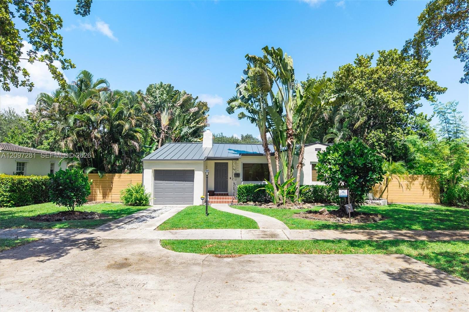Single-story home with a gray metal roof, attached garage, curved driveway, and tropical landscaping.