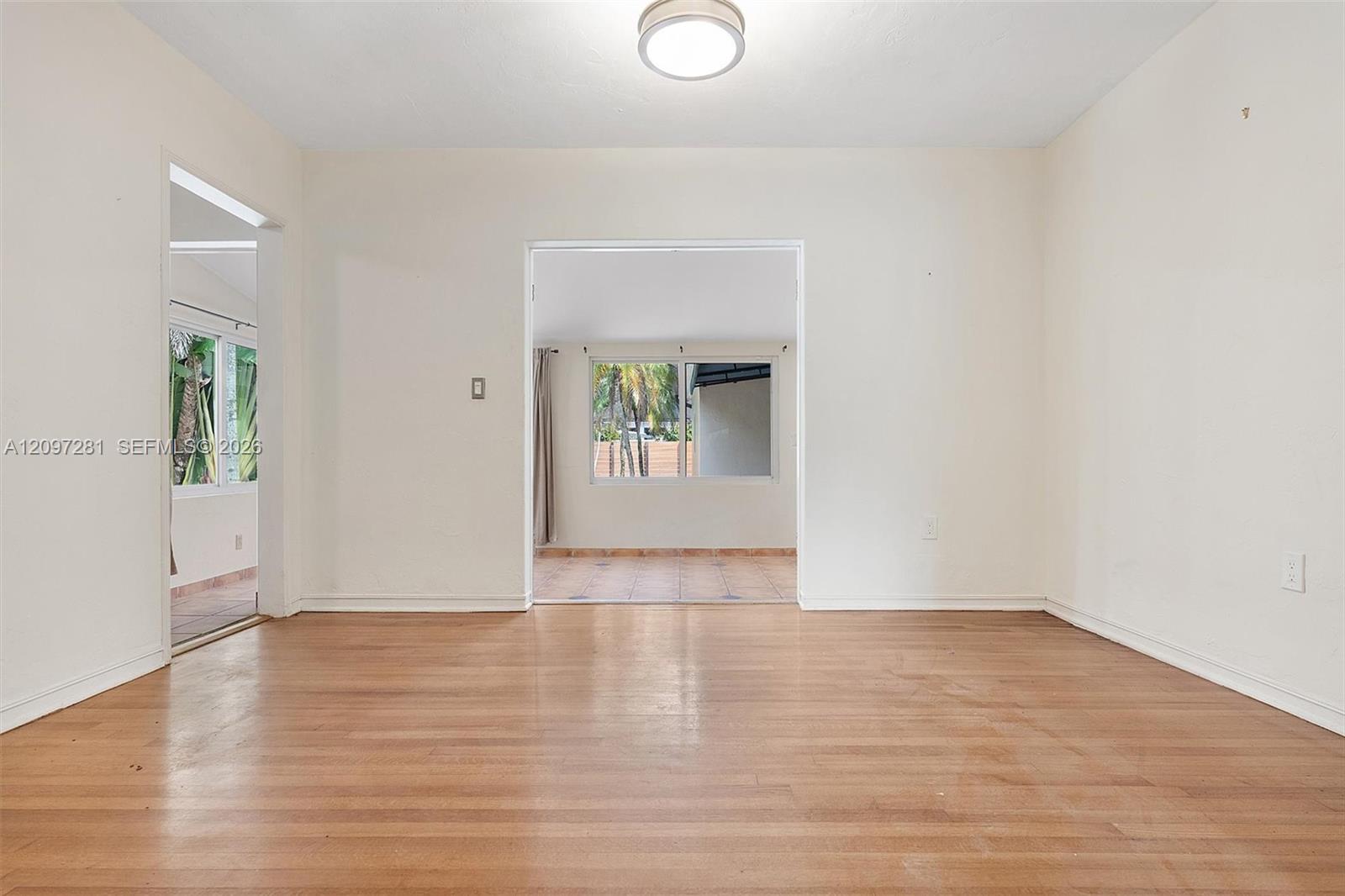 Empty living room with hardwood floors and open doorways to a tiled room with palm views.