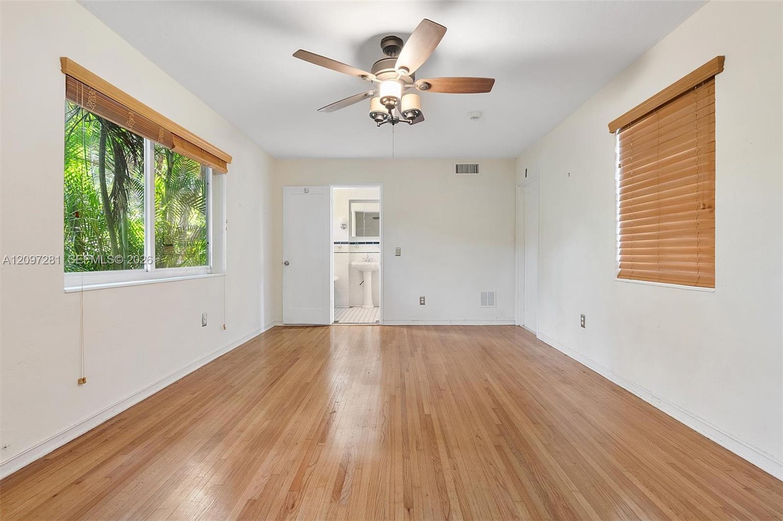 Empty bedroom with hardwood floors, a ceiling fan, and an open doorway to a bathroom.
