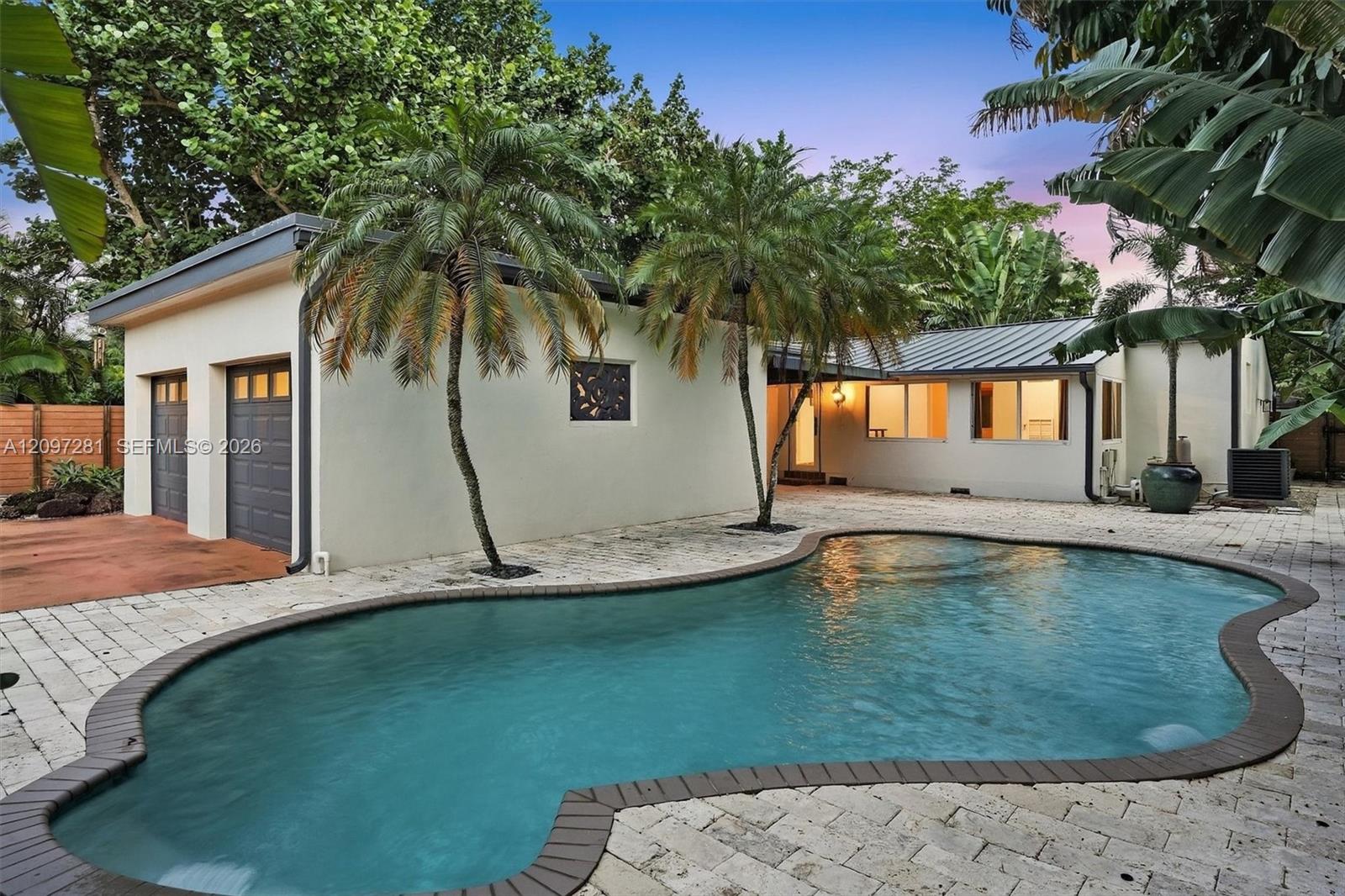 Single-story home with a curved swimming pool, stone patio, and tropical palms at dusk.
