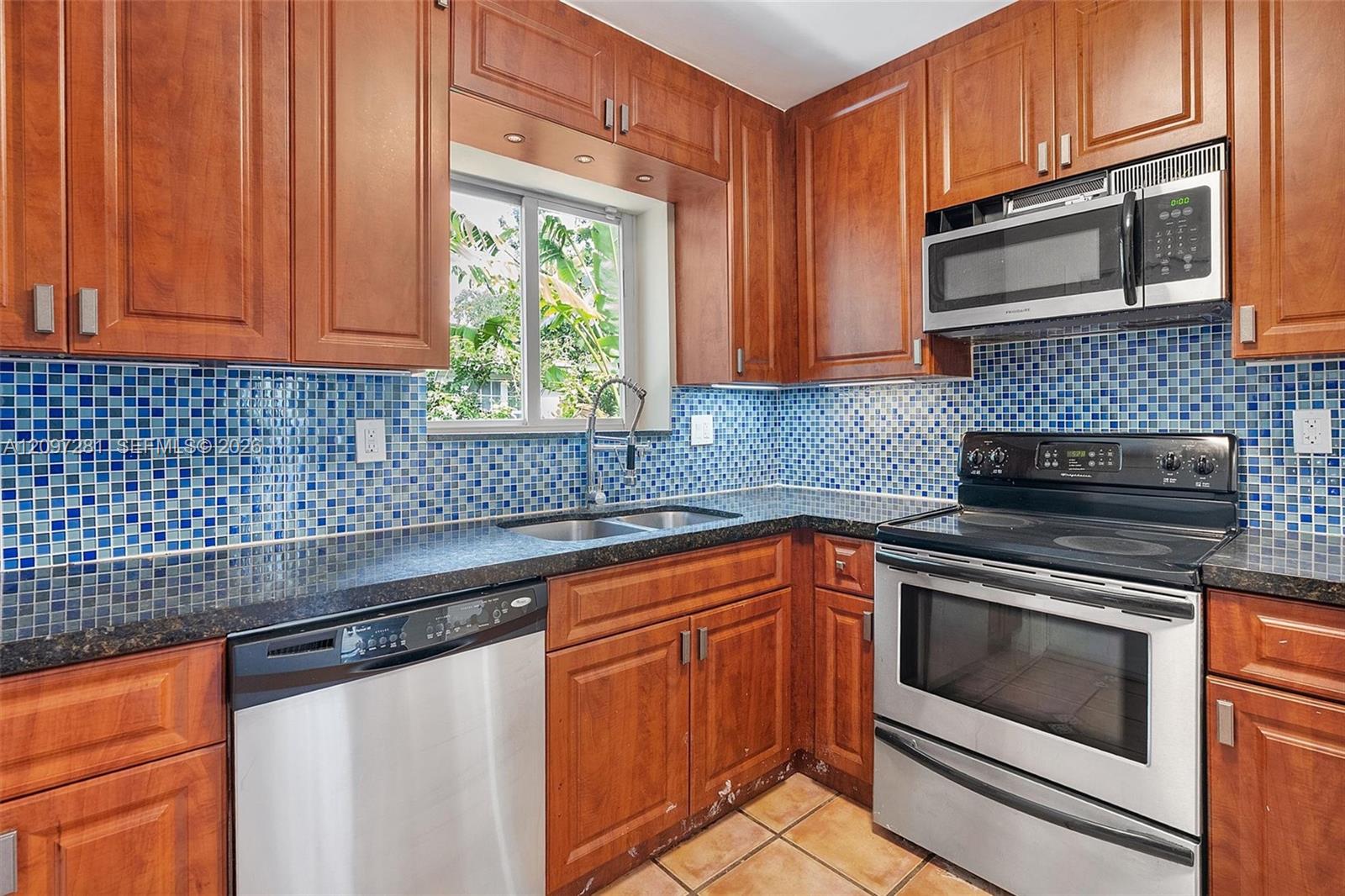 Kitchen with cherry cabinets, blue mosaic backsplash, stainless appliances, and a sink beneath a window.