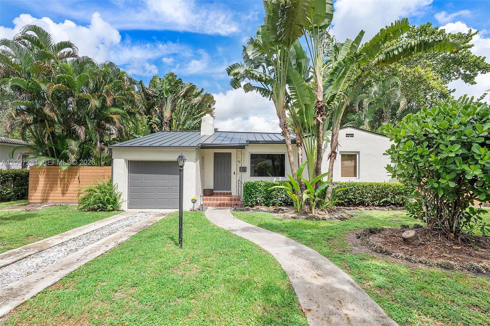 Single-story white house with a gray metal roof, attached garage, and tropical palms in the front yard.