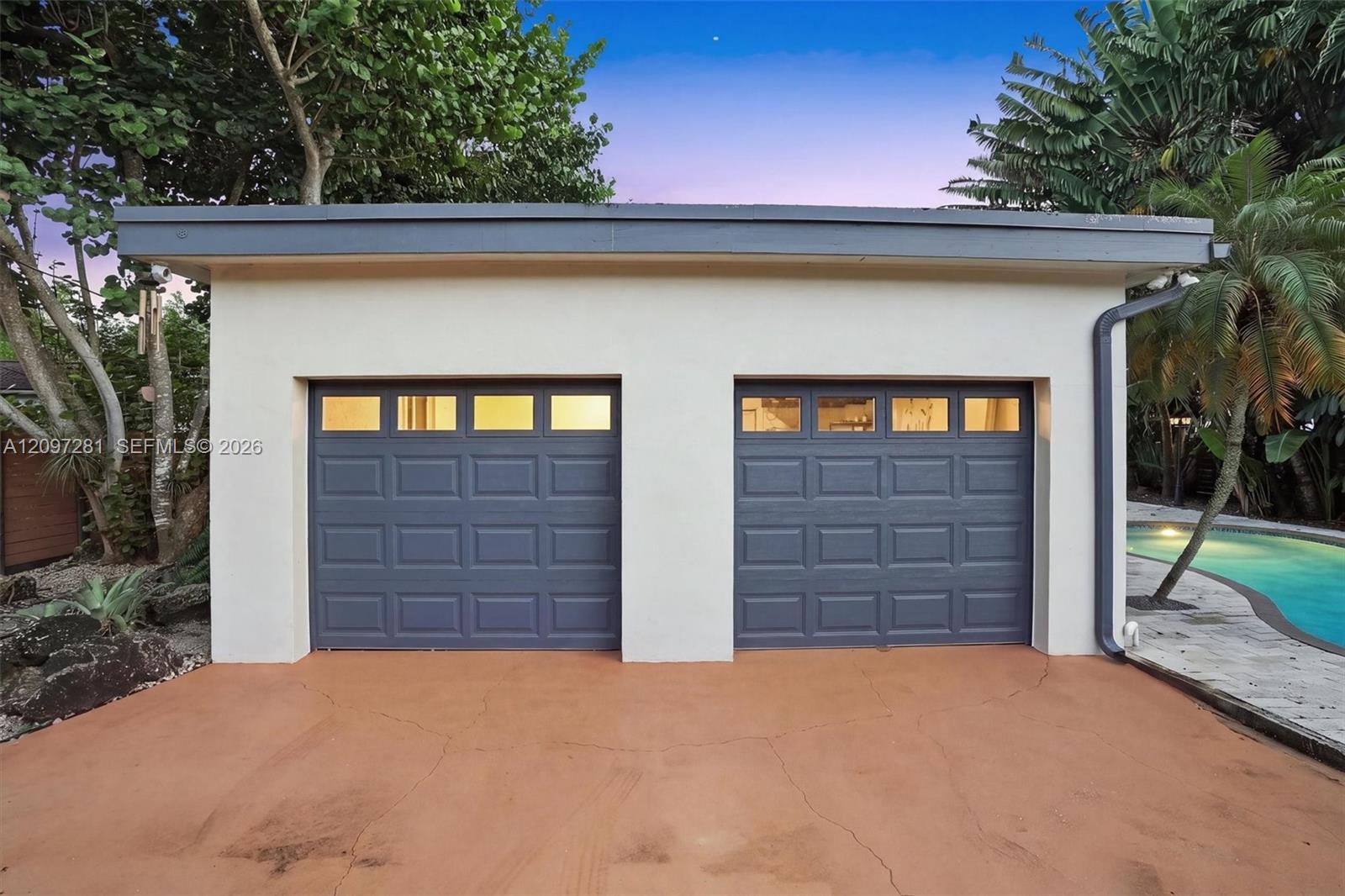 Detached two-car garage with dark blue doors, beside a swimming pool and tropical landscaping.