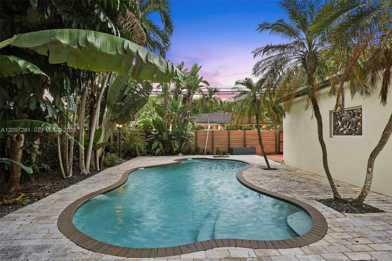 Curving backyard pool surrounded by tropical plants, palm trees, and a wooden privacy fence.