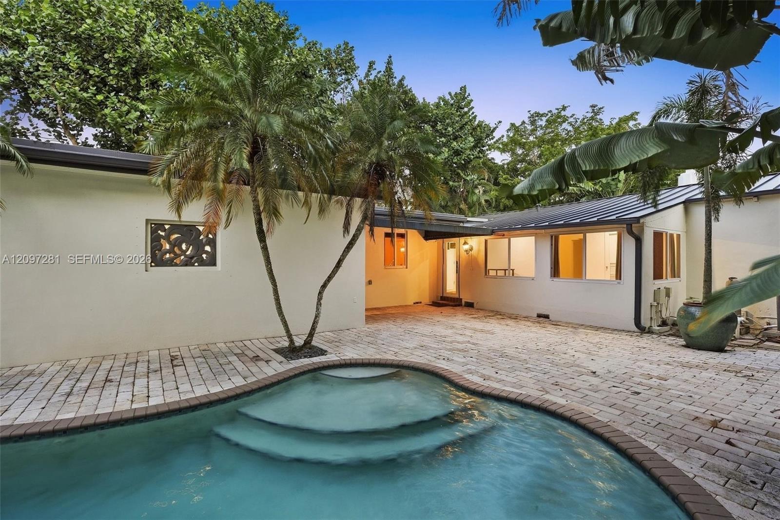 Backyard pool with curved steps beside a white home, paved patio, and tropical palms.