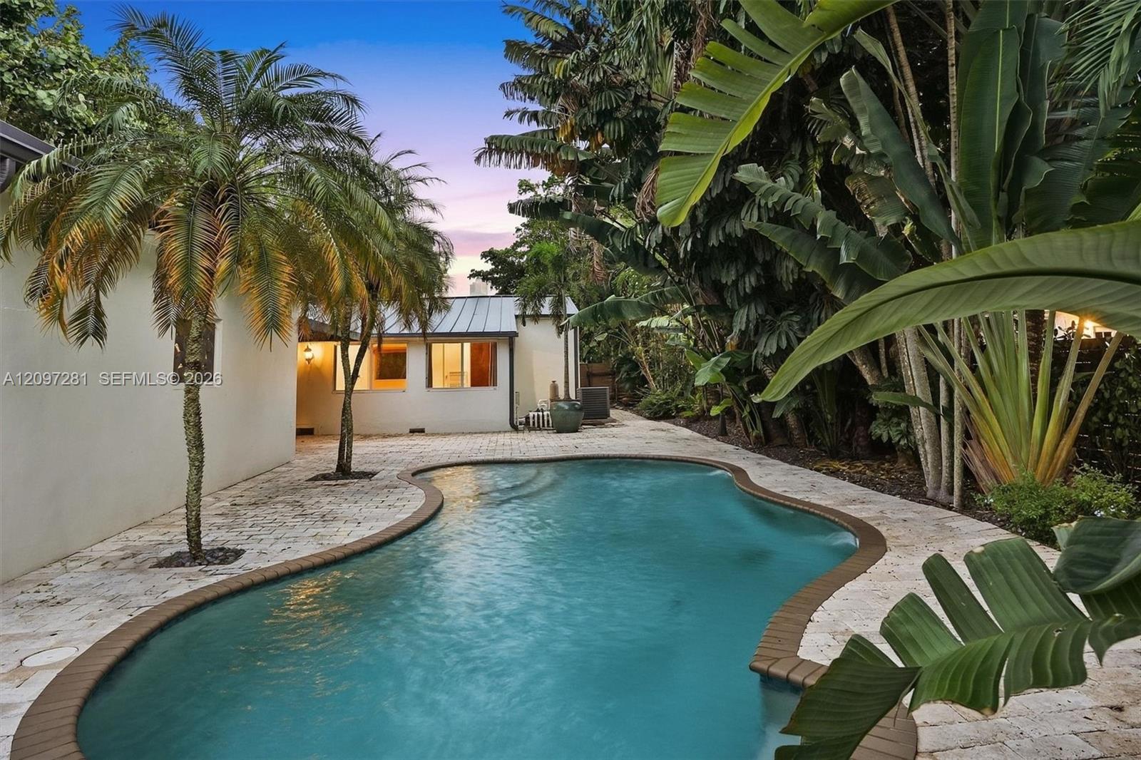 Curved backyard pool beside a white house, framed by dense tropical palms and broad-leaf plants.
